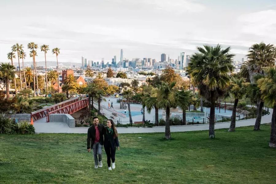 A couple walks toward the camera with Dolores Park and the San Francisco Skyline behind them.