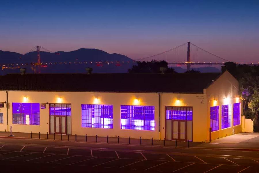The Fort Mason building is lit with a purple interior light at night, with the bridge in the background.