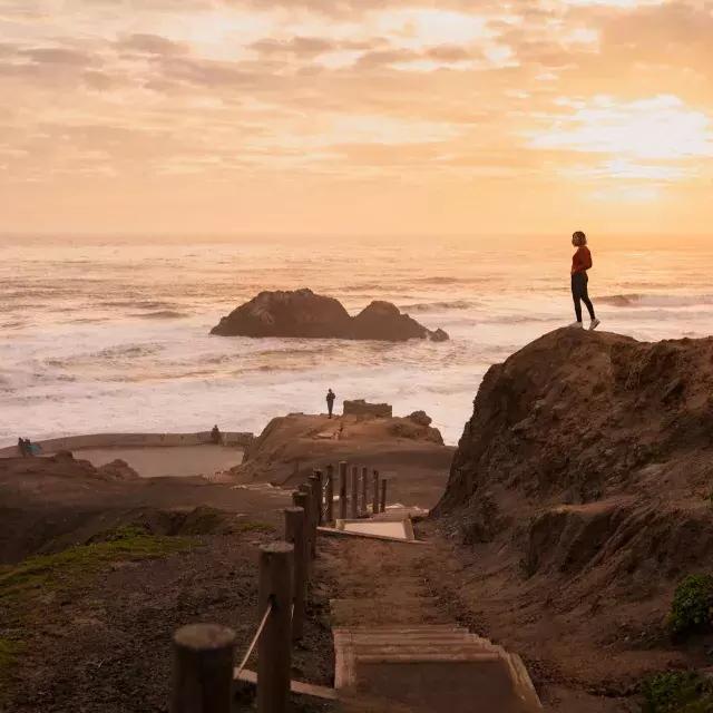 Two people stand on rocks overlooking the ocean at Sutro Baths in San Francisco.